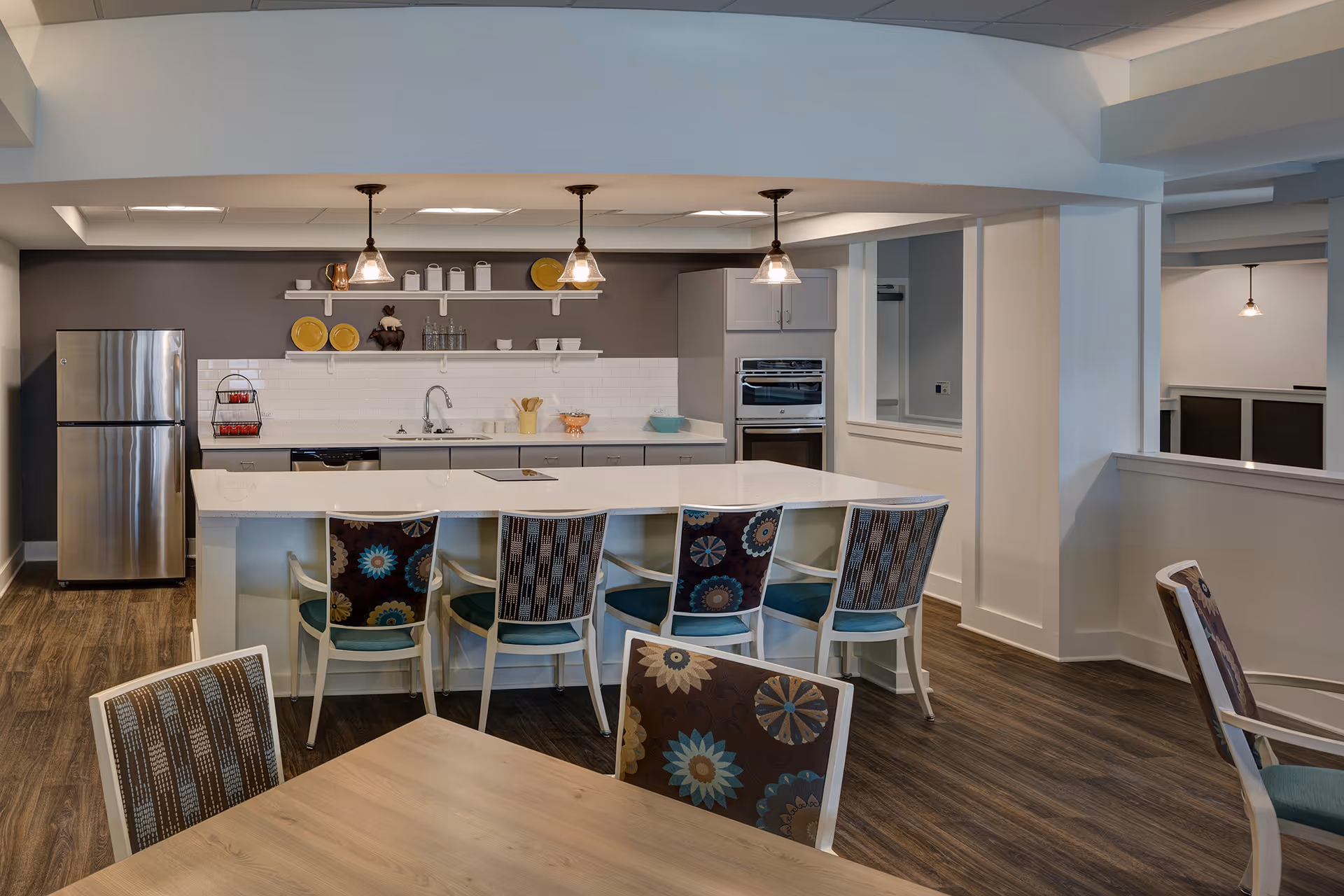 Communal kitchen and dining area with a large white island, patterned chairs, stainless steel refrigerator, pendant lights, and wood flooring.