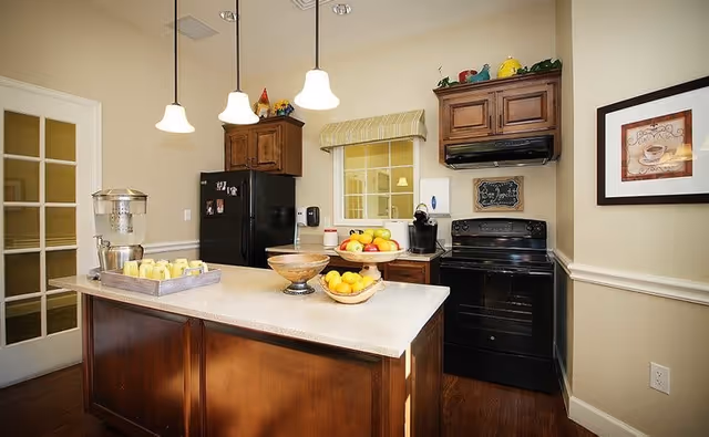 Bright kitchen with a central island, pendant lights, wooden cabinets, black refrigerator and stove, and bowls of fruit on the counter.