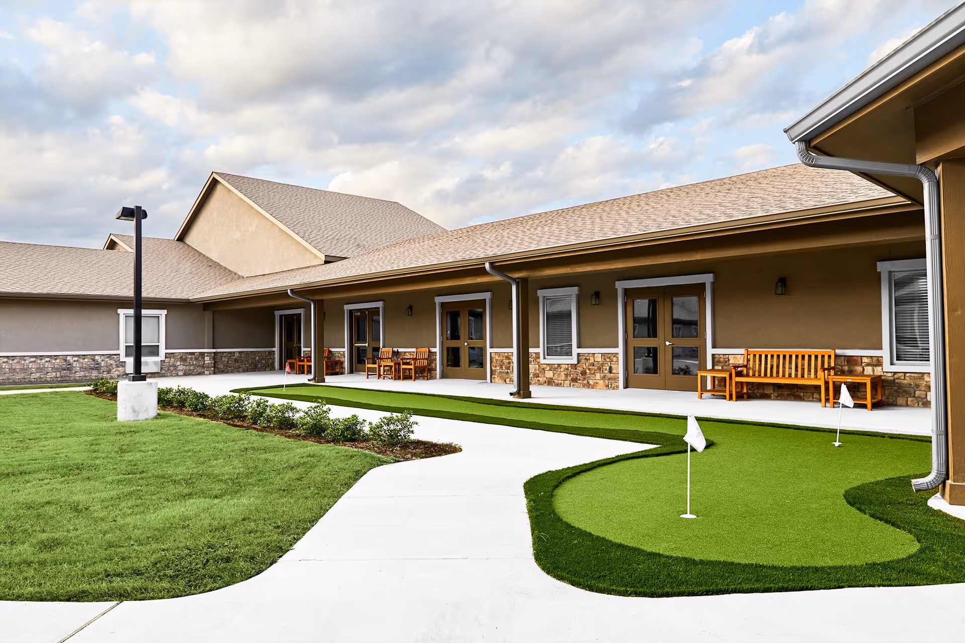 Outdoor area of Le Rêve Rehabilitation and Memory Center featuring a putting green with small flags, a concrete walkway, green grass, and a building with benches and doors under a cloudy sky.