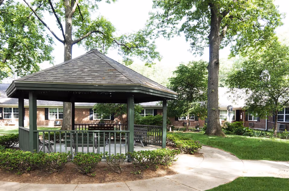 A wooden gazebo in a grassy courtyard surrounded by trees with a brick senior living building in the background.