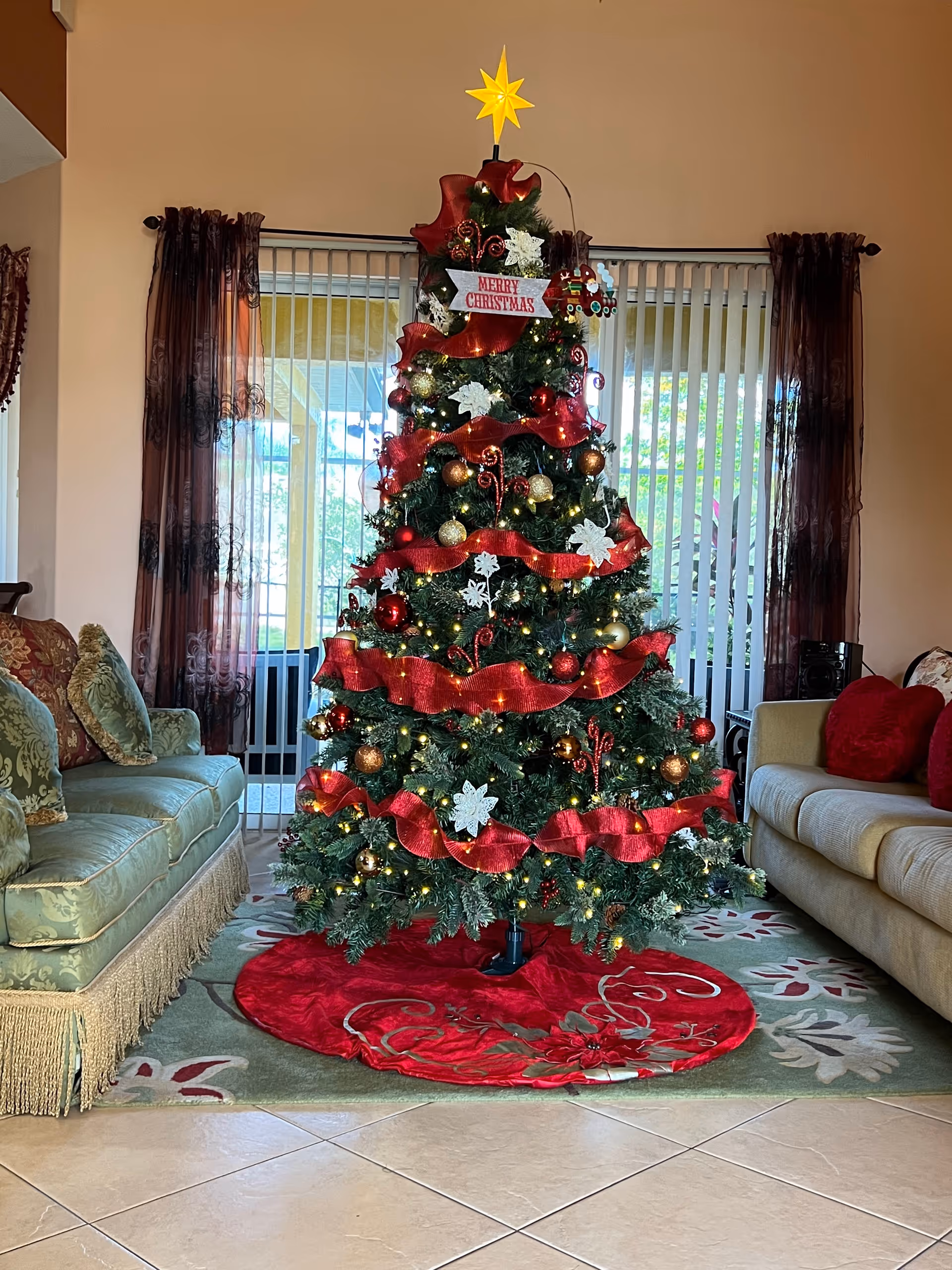 A decorated Christmas tree with red ribbons, gold and red ornaments, white flowers, and a yellow star on top, standing in a living room between two sofas with cushions. The room has tiled floors, a patterned rug, and vertical blinds covering a large window in the background.