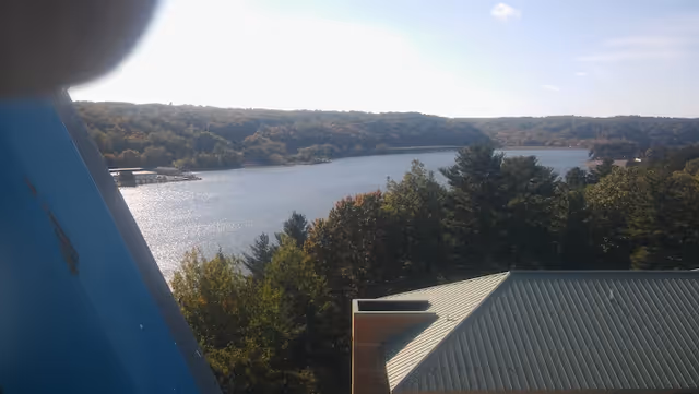 View of a large body of water surrounded by forested hills under a partly cloudy sky, with the roof of a building visible in the foreground.