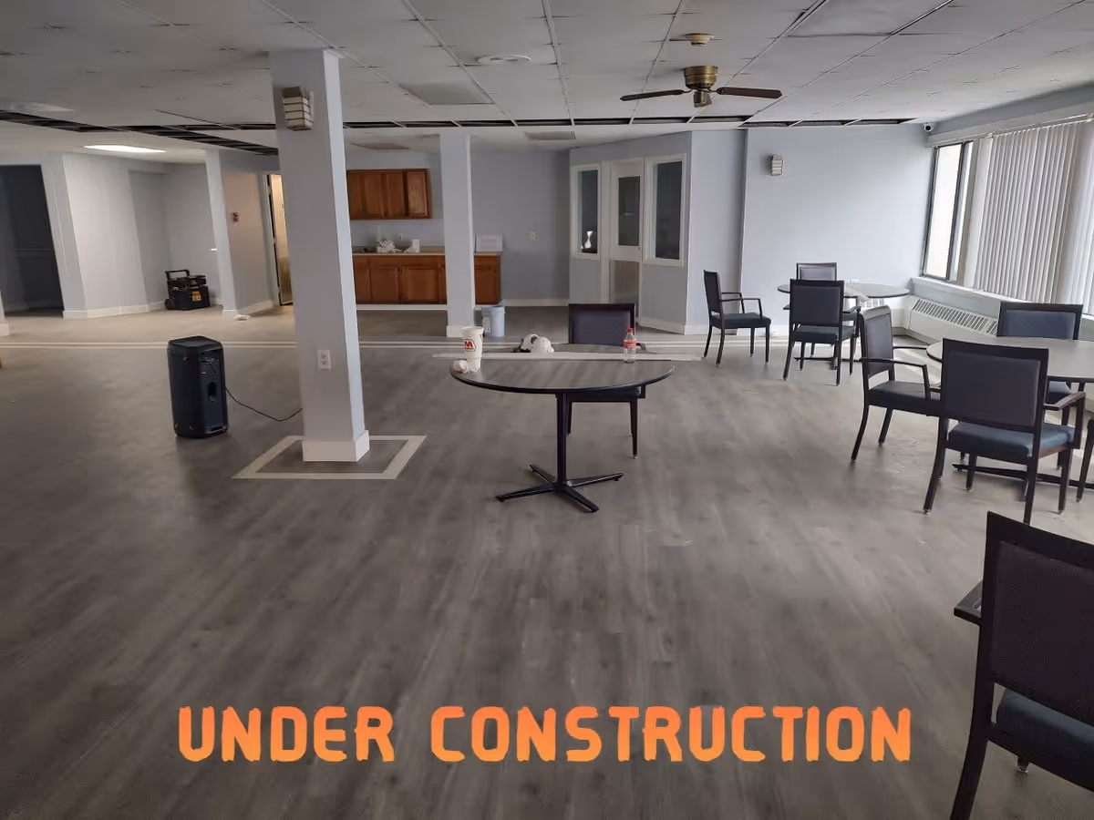 A large interior room under construction with a few tables and chairs scattered around. The ceiling tiles are partially removed, and there are construction materials and equipment visible. The floor is covered with wood-like vinyl flooring. Large windows with vertical blinds allow natural light into the space.