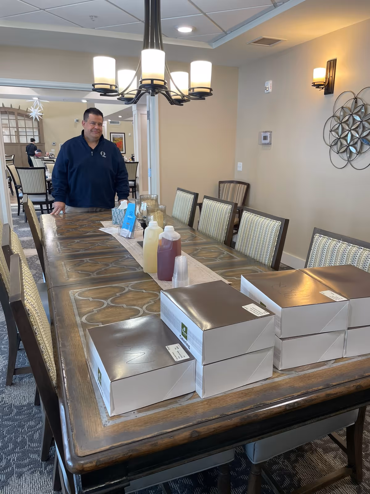 A man standing at the end of a long dining table in a well-lit dining room. The table has several closed white boxes stacked on one end, along with condiment bottles and plastic cups in the center. The room features patterned chairs, a chandelier overhead, wall sconces, and decorative wall art.