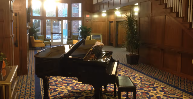 Grand piano in a wood-paneled senior living lobby with patterned carpet, potted plants and seating by large windows.