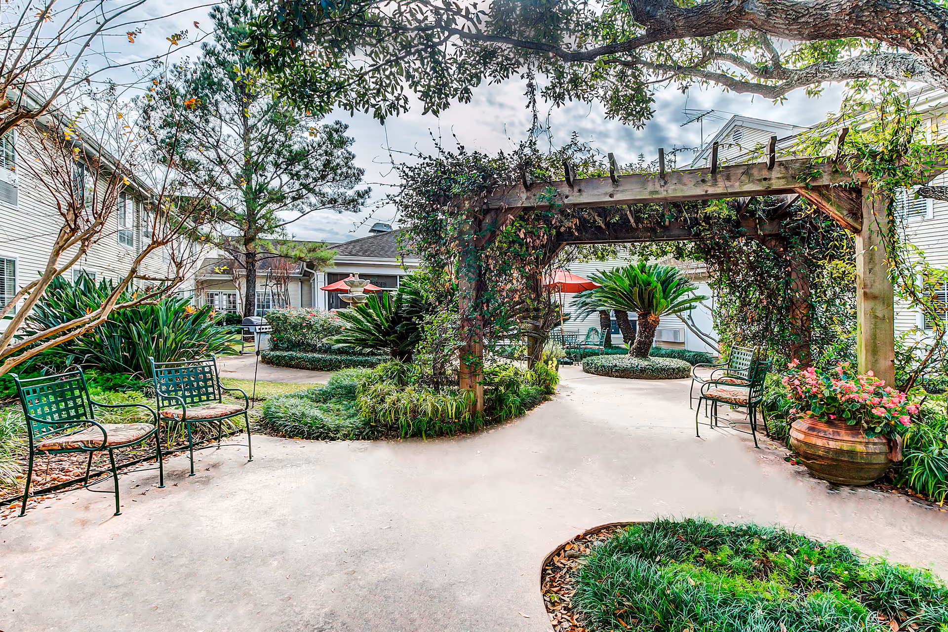 Outdoor garden area at The Gardens of Bellaire featuring a wooden pergola covered with climbing plants, green metal benches with cushions, various shrubs and trees, potted flowers, and a concrete pathway winding through the landscaped space. Residential buildings surround the garden area.