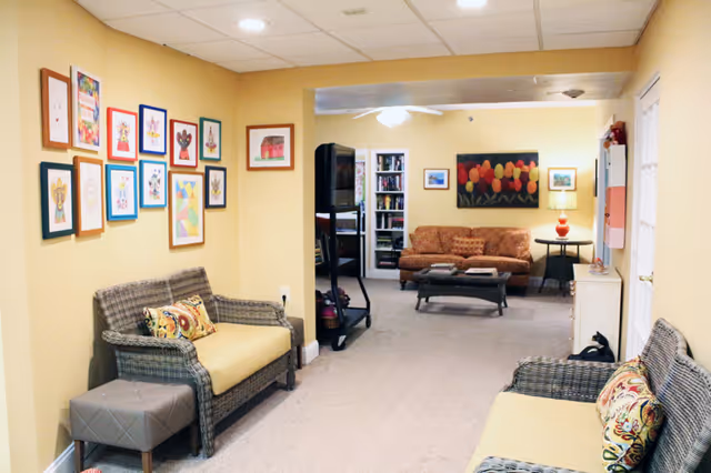 A cozy living room area in an assisted living facility with yellow walls and carpeted floor. The room features two wicker sofas with yellow cushions and patterned pillows, a small gray ottoman, a coffee table, and a brown patterned couch in the background. The walls are decorated with colorful framed artwork, and there is a bookshelf filled with books. A lamp with a red base is on a side table next to the couch, and a ceiling fan is visible above.