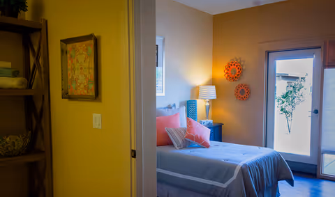 A cozy bedroom with a neatly made bed, colorful pillows, bedside lamp and wall decor, with a glass door leading outside.
