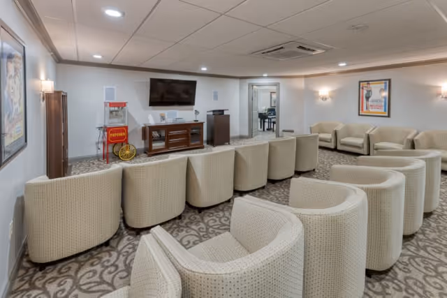 A spacious living room area with multiple beige upholstered armchairs arranged in rows facing a wall-mounted flat screen TV. The room has patterned carpet flooring, white walls with framed artwork, and recessed ceiling lights. A popcorn machine is placed near the TV stand, and there is an open doorway leading to another room with a piano visible.