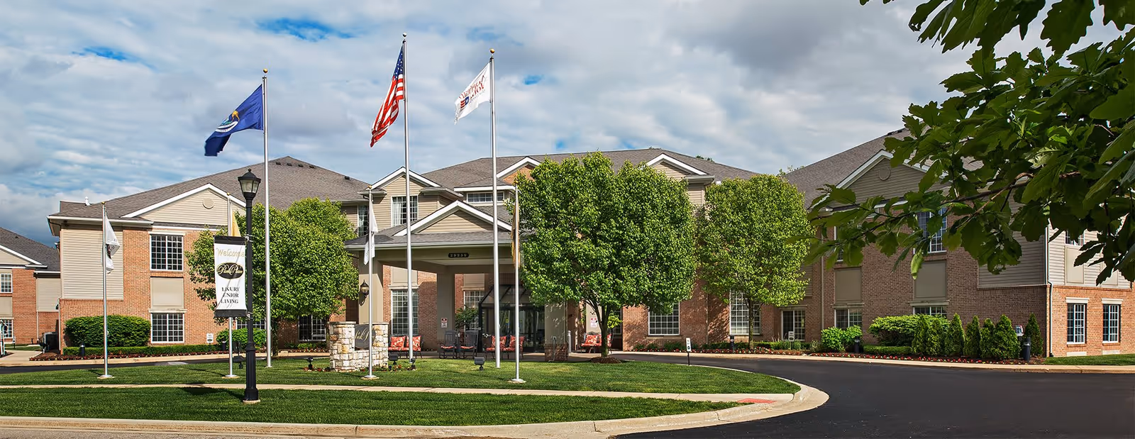 Exterior view of a senior living facility named American House Park Place with a well-maintained lawn, several trees, and multiple flagpoles displaying flags including the American flag. The building is two stories with a brick and siding facade under a partly cloudy sky.