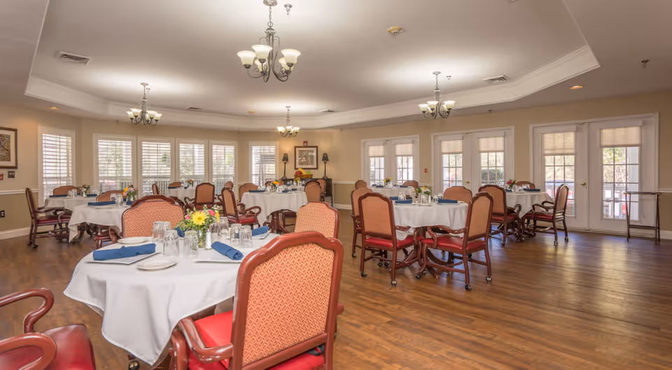 Spacious dining room with round tables covered in white tablecloths, place settings, and upholstered chairs.