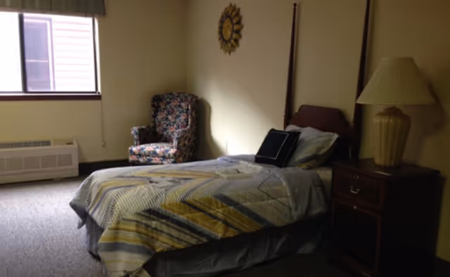 Small bedroom with a single bed, wooden nightstand and lamp, a floral armchair by a window and a sunburst wall decoration.
