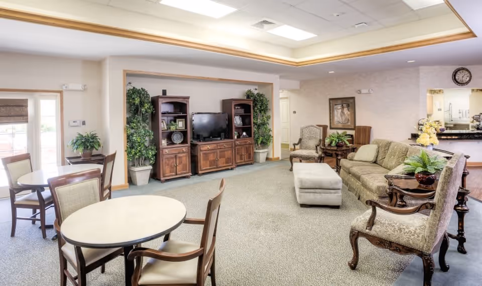 A bright and spacious living room area in a senior living facility featuring a green upholstered sofa, two armchairs, a cushioned ottoman, and wooden side tables with plants. There is a TV on a wooden entertainment center flanked by two tall plants. In the foreground, there are round tables with chairs for dining or socializing. The room has carpeted flooring, a clock on the wall, and a pass-through window to the kitchen area.
