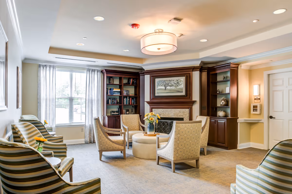 Well-lit senior living common room with upholstered chairs arranged around a central ottoman, a fireplace and built-in bookshelves.