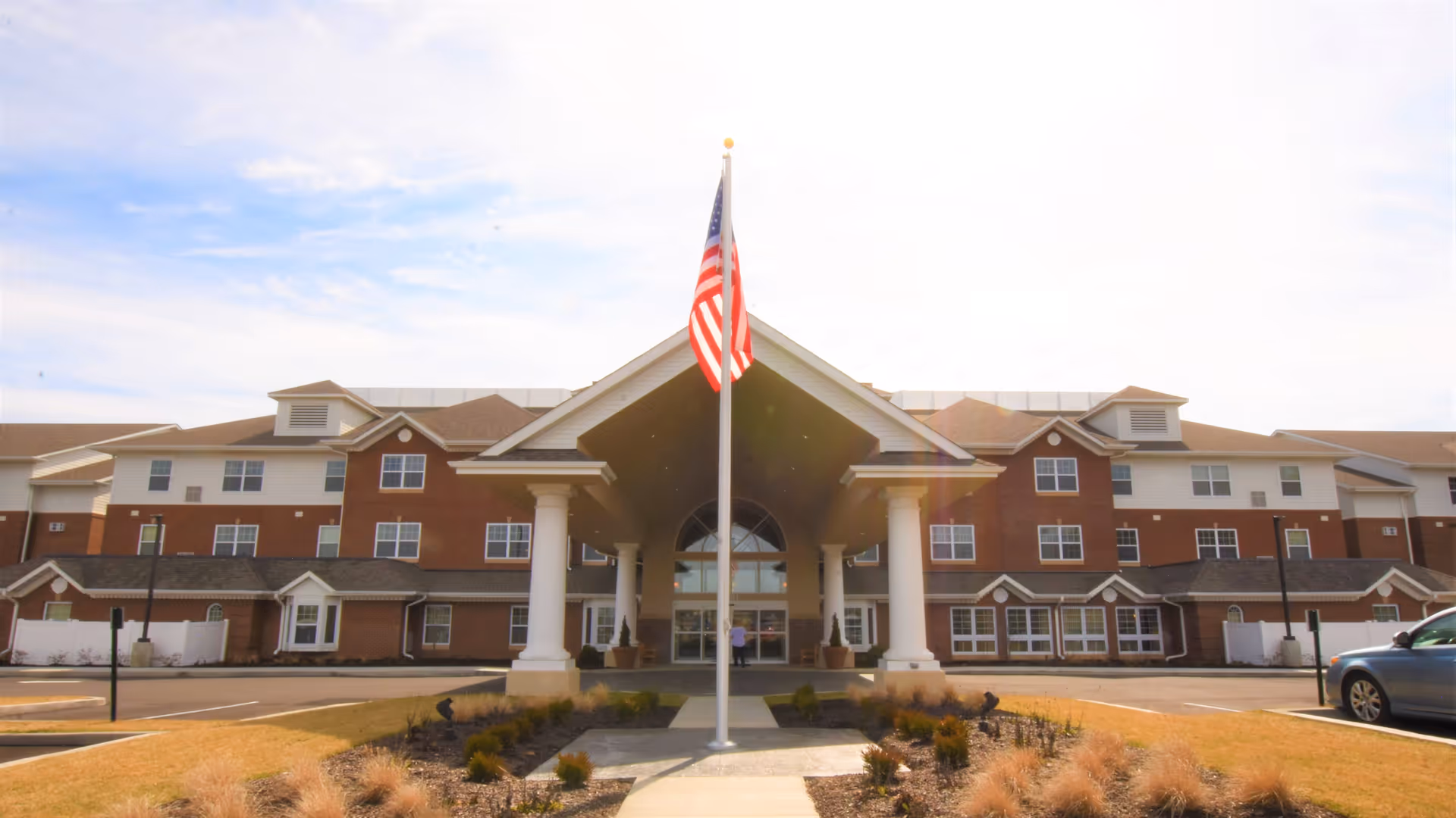 Front exterior view of Chartiers Bend Retirement Resort, a large multi-story building with a covered entrance supported by white columns. An American flag is prominently displayed on a flagpole in front of the entrance. The sky is partly cloudy and there are some parked cars and landscaped areas around the building.