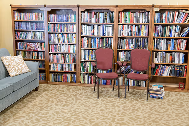 A cozy library-style room with tall wooden bookshelves, two red chairs, a stack of books on the floor, and a sofa with a patterned pillow.