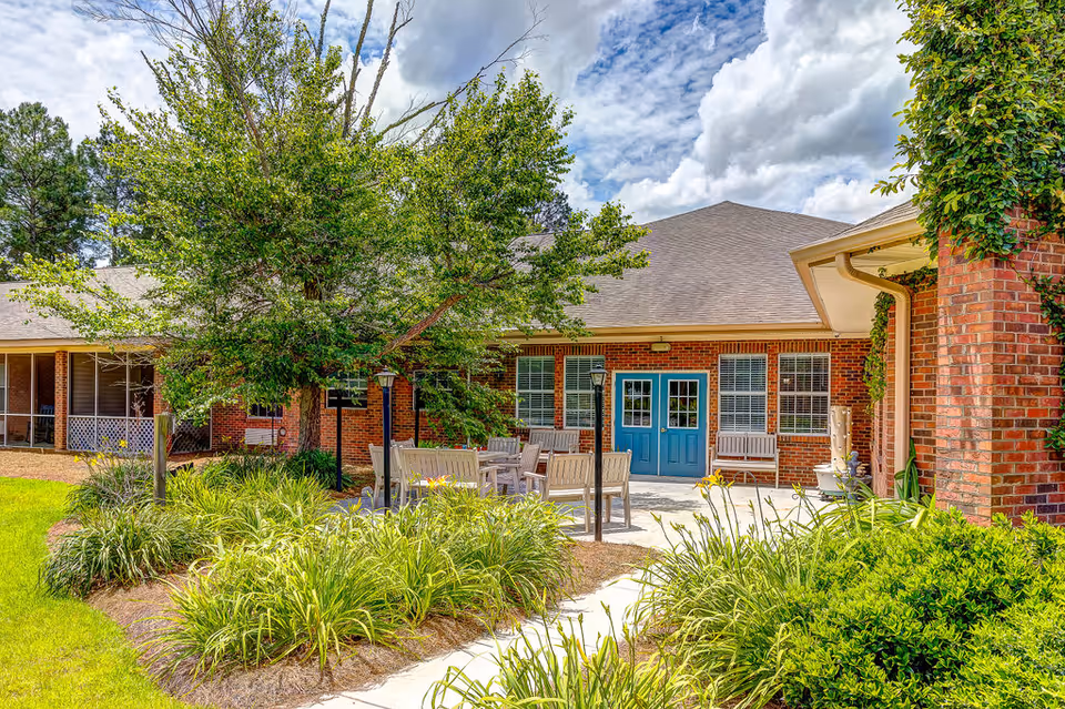 Outdoor patio area at Legacy Village at Park Regency featuring a brick building with blue double doors, surrounded by green plants and trees under a partly cloudy sky. There are several wooden chairs and tables on the patio.
