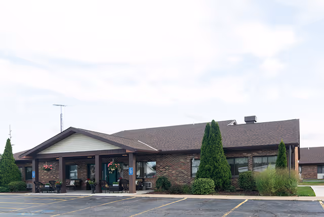 Exterior view of a single-story brick building with a covered entrance supported by wooden beams. There are several potted plants and hanging flower baskets near the entrance. The building is surrounded by neatly trimmed bushes and tall evergreen trees. In front of the building is a parking lot with marked parking spaces under a partly cloudy sky.