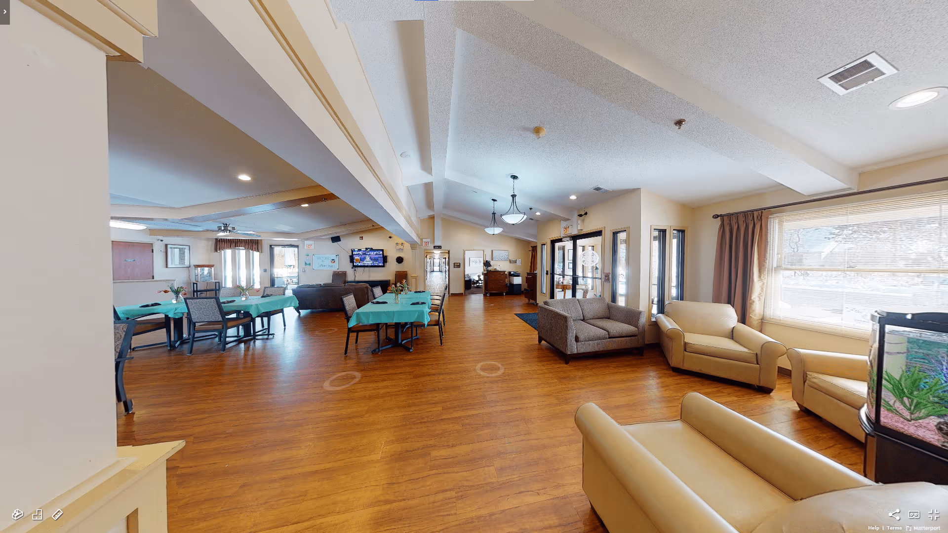 A spacious senior living common area with wooden flooring, beige walls, and a high ceiling with recessed lighting and hanging lamps. The room features several tables covered with green tablecloths and chairs, a seating area with beige and gray armchairs and sofas near large windows with blinds, and a fish tank in the corner. A television is mounted on the far wall above a dark sofa, and there are exit signs and doors visible in the background.
