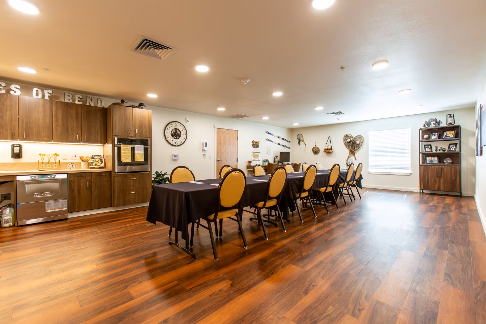 A spacious room with a long table covered with a black tablecloth and surrounded by yellow cushioned chairs. The room features wooden flooring, a kitchenette with wooden cabinets, a dishwasher, and an oven. The walls are decorated with various items including a clock, vintage skis, and snowshoes. A window with blinds and a wooden cabinet with framed photos and decorations are visible at the far end.
