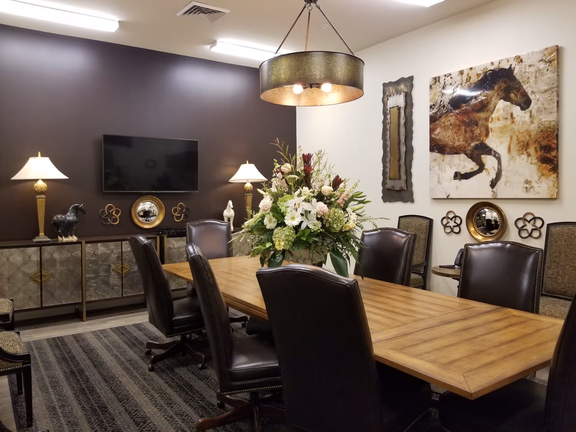 A well-decorated meeting or dining room with a long wooden table surrounded by black leather chairs. The room features a large floral centerpiece on the table, two table lamps on a sideboard against a dark brown accent wall, a flat-screen TV mounted on the wall, and decorative horse-themed artwork and mirrors on the walls. The room has a modern chandelier hanging from the ceiling and a patterned carpet on the floor.
