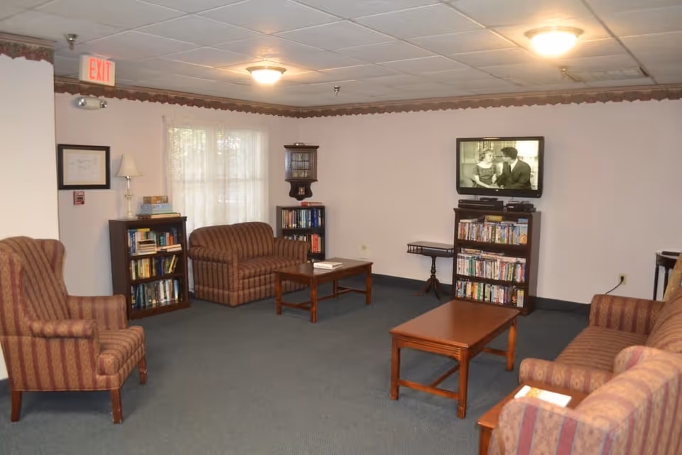 A cozy senior living common area with striped upholstered armchairs and sofas arranged around wooden coffee tables. There are bookshelves filled with books and DVDs, a wall-mounted TV showing a black-and-white movie, and a window with sheer curtains letting in natural light. The room has a carpeted floor and a drop ceiling with ceiling lights.