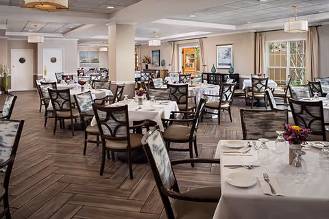 A spacious dining room with multiple tables covered in white tablecloths, each set with plates, glasses, and silverware. The room features wooden herringbone-patterned flooring, cushioned chairs with patterned backs, large windows with beige curtains, and soft overhead lighting. Decorative elements include framed artwork on the walls and floral centerpieces on the tables.