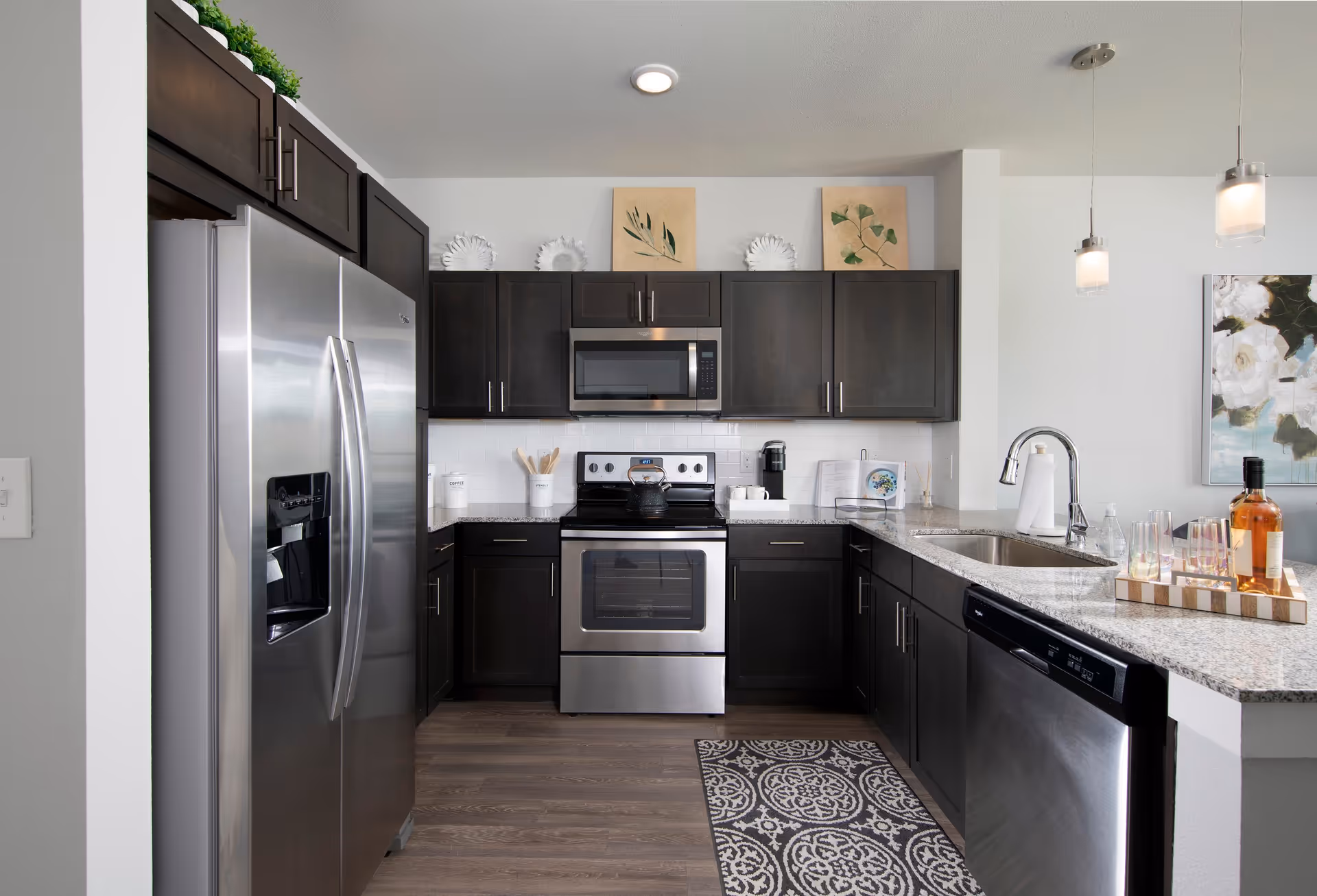 Modern kitchen with dark wood cabinets, stainless steel refrigerator, oven, microwave, and dishwasher. Granite countertops with a sink and faucet, decorative plates and artwork above the cabinets, and pendant lights hanging from the ceiling. A patterned rug is on the wooden floor.