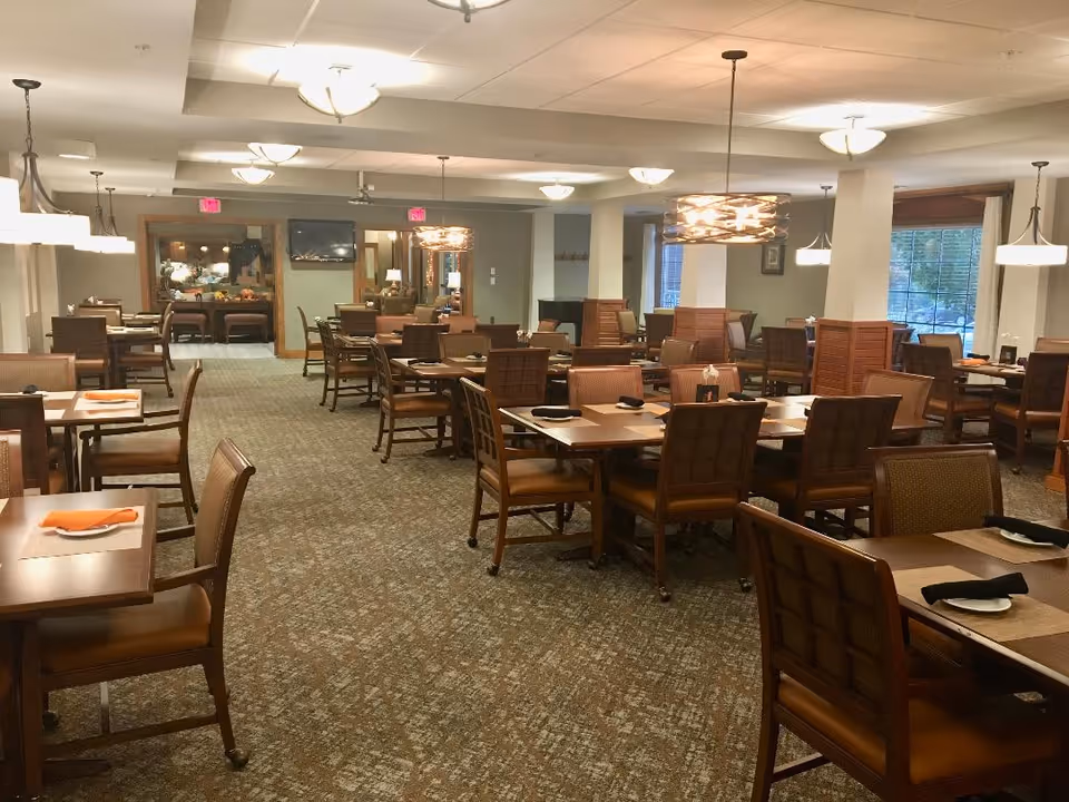 A spacious dining room with multiple wooden tables and chairs arranged neatly. Each table is set with plates and napkins, some orange and some black. The room is well-lit with ceiling lights and pendant lamps. Large windows on the right side allow natural light to enter, and there are decorative elements such as framed pictures on the walls.