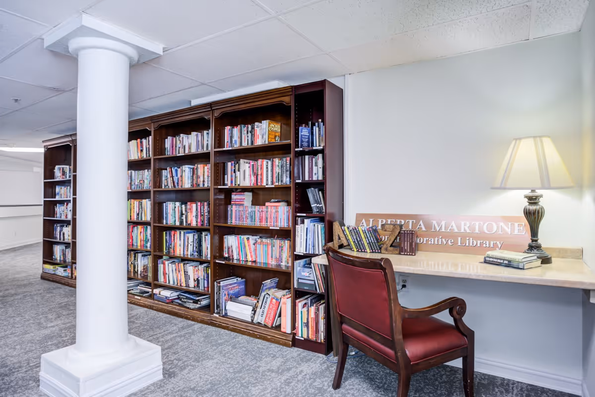 Interior view of a library area with tall wooden bookshelves filled with books along the wall. A wooden chair with red upholstery is placed in front of a small desk with a table lamp and a sign that reads 'ALBERTA MARTONE Memorial Library'. The floor is carpeted and a white column is visible in the foreground.
