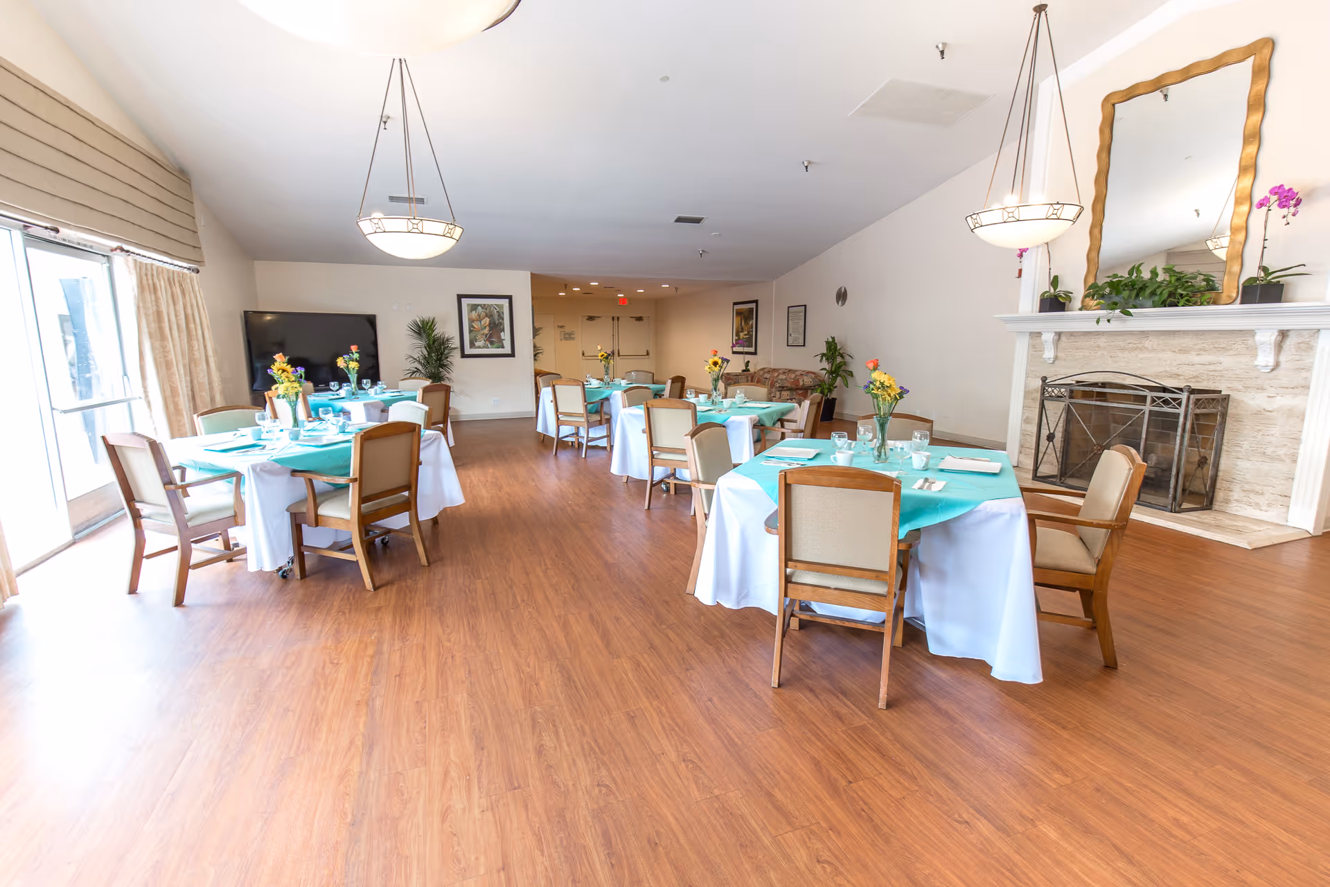 A bright dining room in a senior living facility with round tables covered in white and turquoise tablecloths, each set with glassware, plates, and floral centerpieces. The room features wooden flooring, a fireplace with a large mirror above it, framed artwork on the walls, and large windows with curtains allowing natural light to enter.
