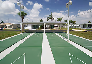 Outdoor shuffleboard courts with green playing surfaces and white markings, surrounded by grass and palm trees, under a partly cloudy sky. Residential buildings are visible in the background.