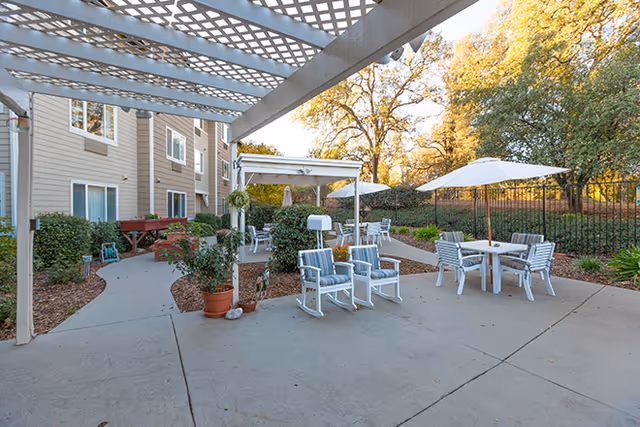 Outdoor patio area at a senior living facility with white rocking chairs, tables with umbrellas, potted plants, and a pergola providing partial shade. The building exterior and landscaped garden with trees are visible in the background.