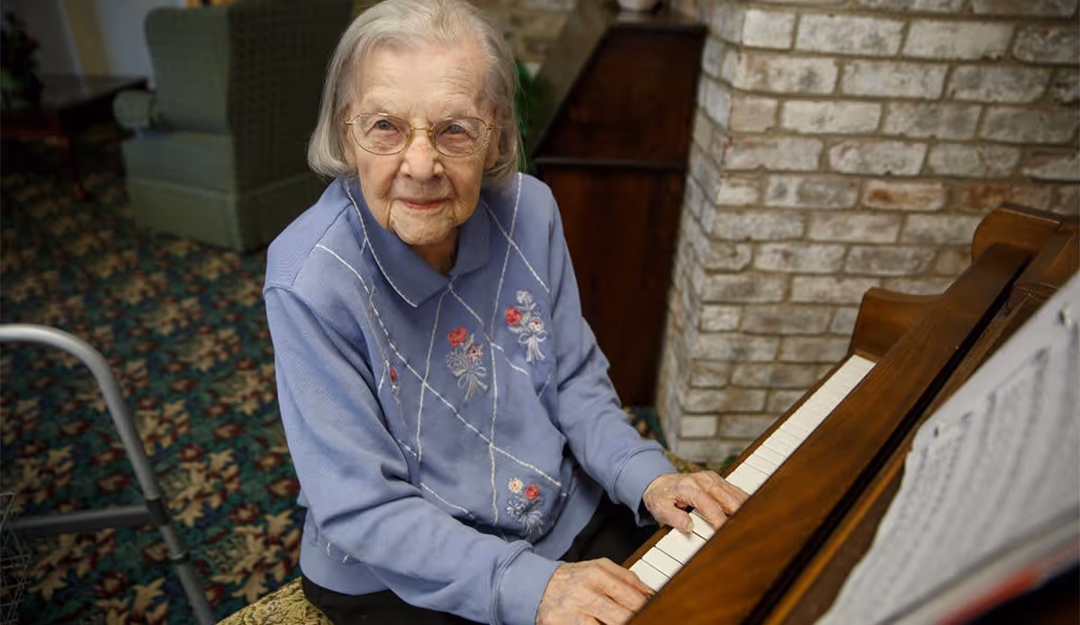 An elderly woman wearing glasses and a blue sweater with floral embroidery is seated at a piano, playing music. She is looking up towards the camera with a gentle expression. The background shows a patterned carpet, a green armchair, a walker, and a brick wall.