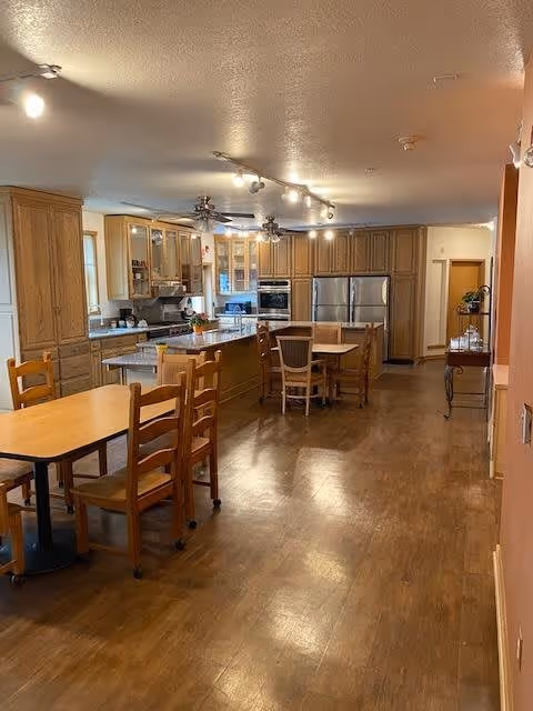 A spacious kitchen area with wooden cabinets, stainless steel refrigerator, built-in ovens, and a kitchen island with chairs. There are multiple wooden tables and chairs arranged in the room, with ceiling lights and ceiling fans providing illumination.