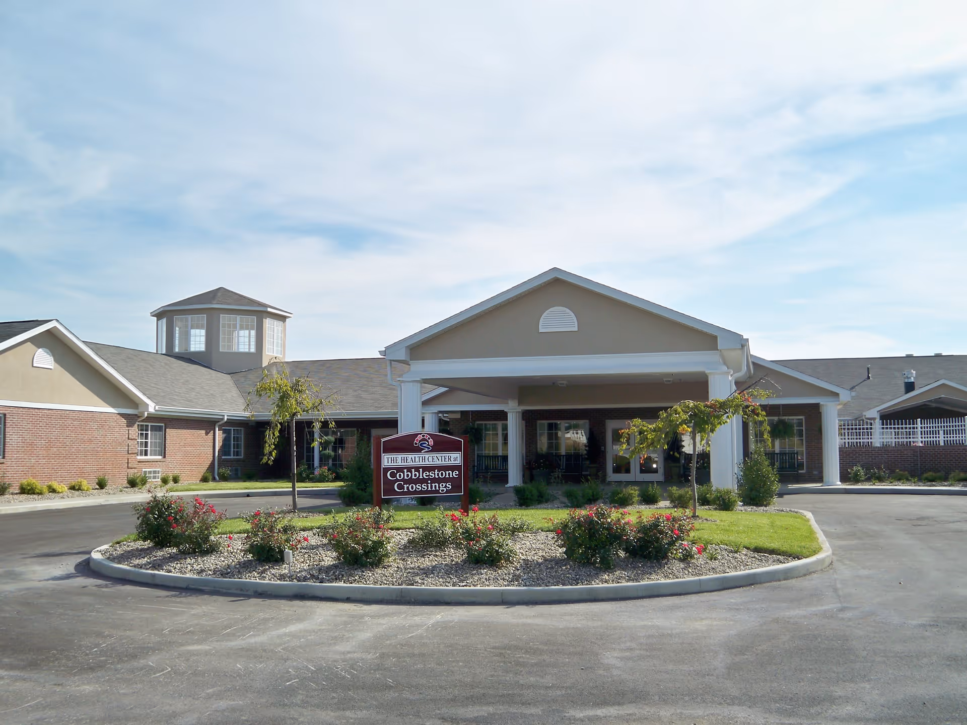Front exterior view of Cobblestone Crossings health center building with a circular driveway, landscaped garden with flowers and small trees, and a sign displaying the facility name.