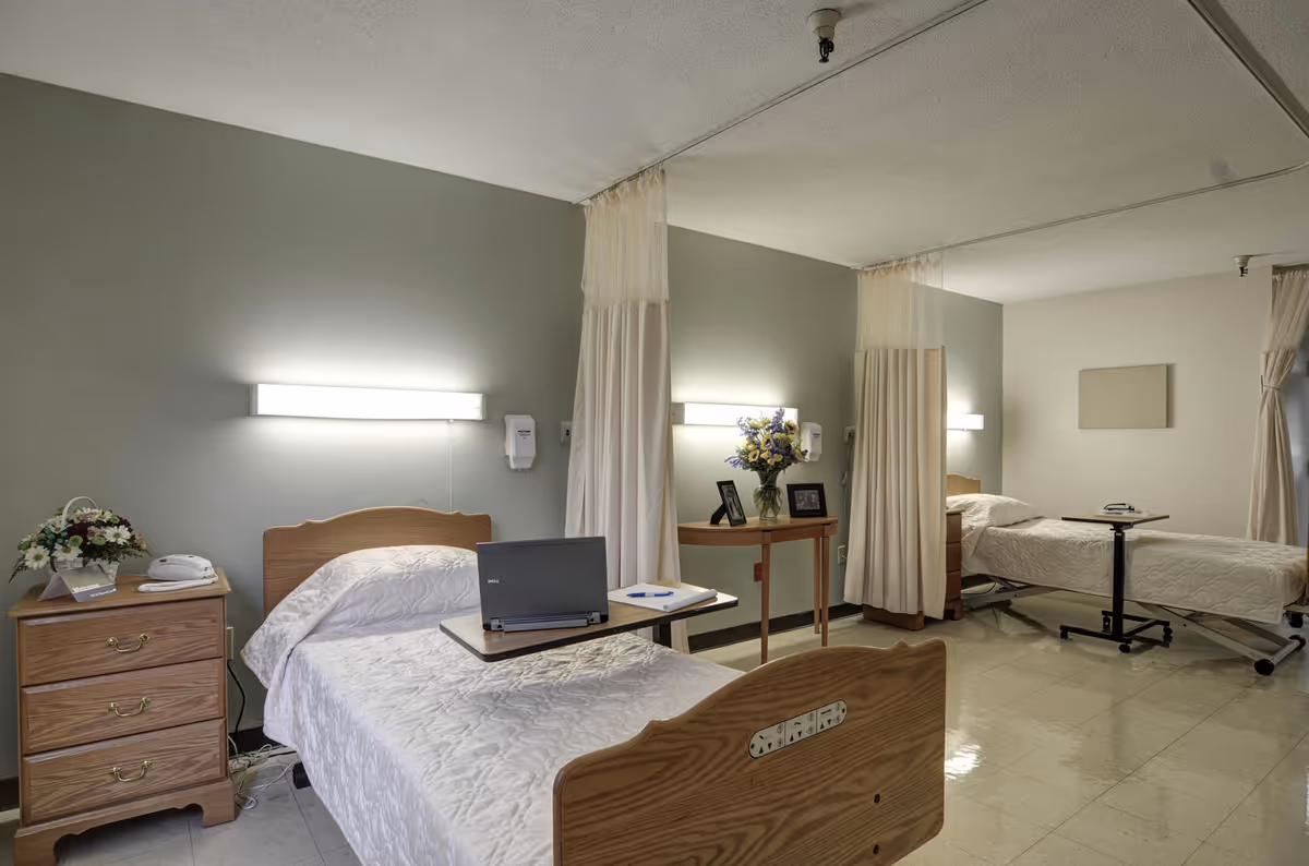 A shared patient room in a healthcare facility with two hospital beds separated by privacy curtains. Each bed has a bedside table with flowers and a telephone. One bed has a laptop and notebook on an overbed table. The room has soft lighting and a clean, simple design.