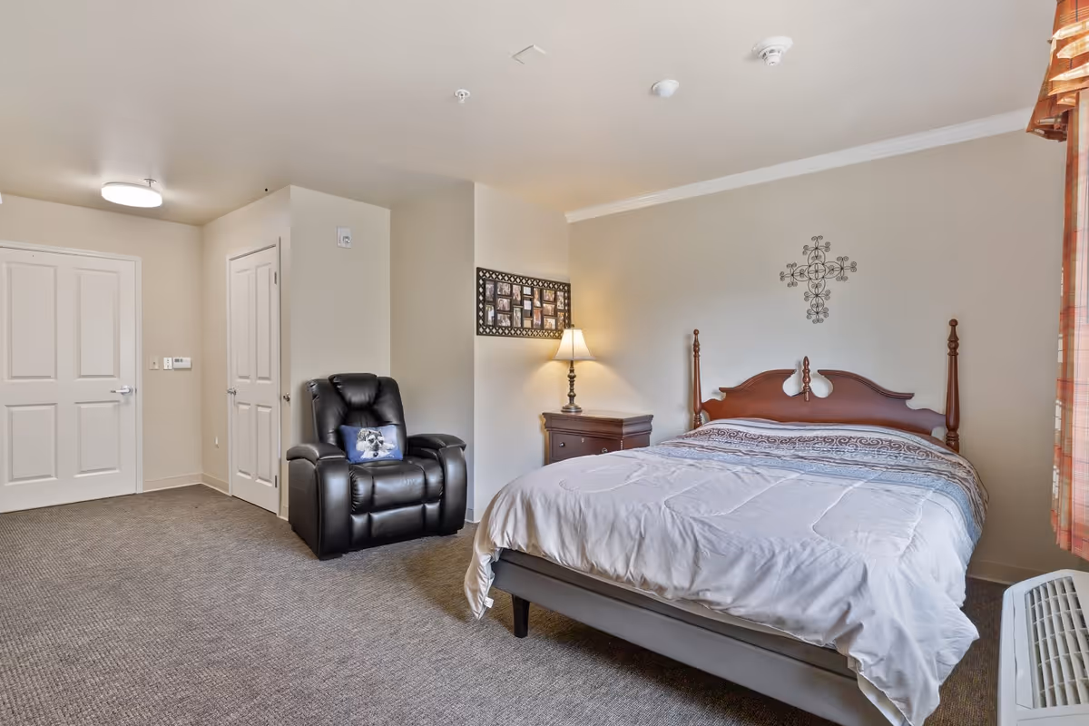 A bedroom with a large bed featuring a wooden headboard, a nightstand with a lamp, a black recliner chair with a decorative pillow, a wall decoration above the bed, and two closed white doors. The room has beige walls, carpeted floor, and a window with plaid curtains partially visible on the right.