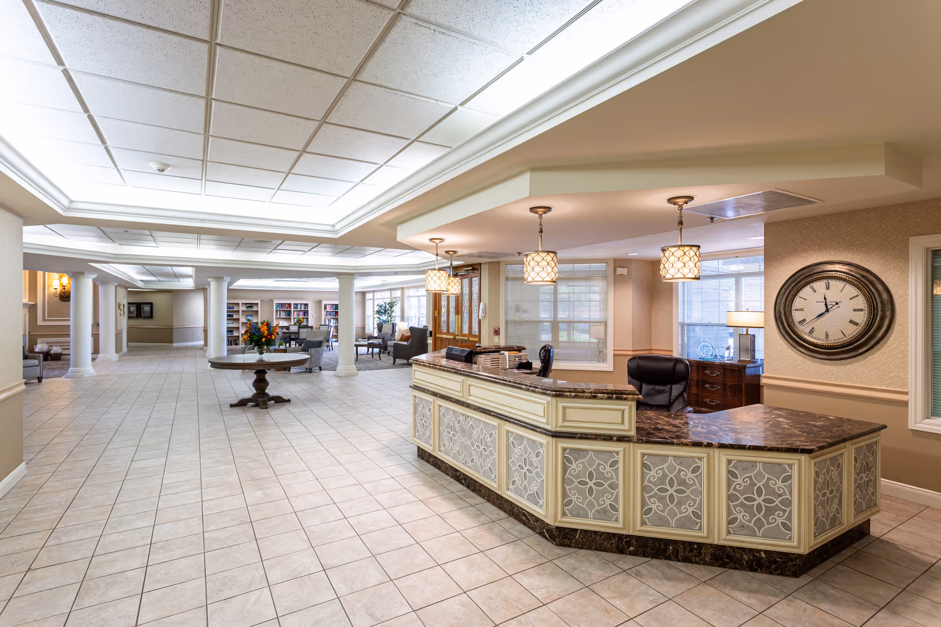 Spacious and well-lit reception area with a large decorative front desk featuring a marble countertop and intricate panel designs. Behind the desk is a comfortable office chair and a wall clock. The area has tiled flooring, columns, and a round table with a flower arrangement in the middle. In the background, there are seating areas with chairs and bookshelves near large windows allowing natural light.