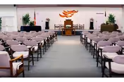A large room set up with rows of chairs facing a stage area with a podium and decorative wall art. The chairs have light-colored cushions and wooden armrests. There are plants on either side of the stage and the room has a carpeted floor.