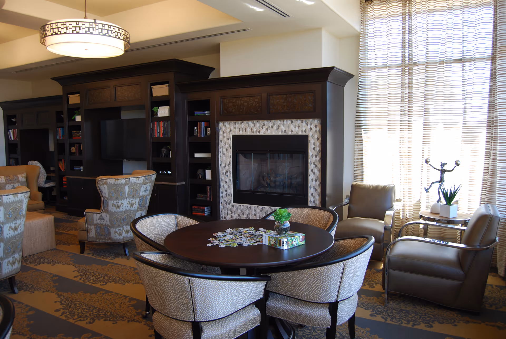 A cozy living room area with a round table surrounded by four upholstered chairs, a fireplace with a decorative tile surround, built-in dark wood shelves filled with books and decor, two leather armchairs near a window with sheer curtains, and a small side table with a plant and a decorative sculpture.