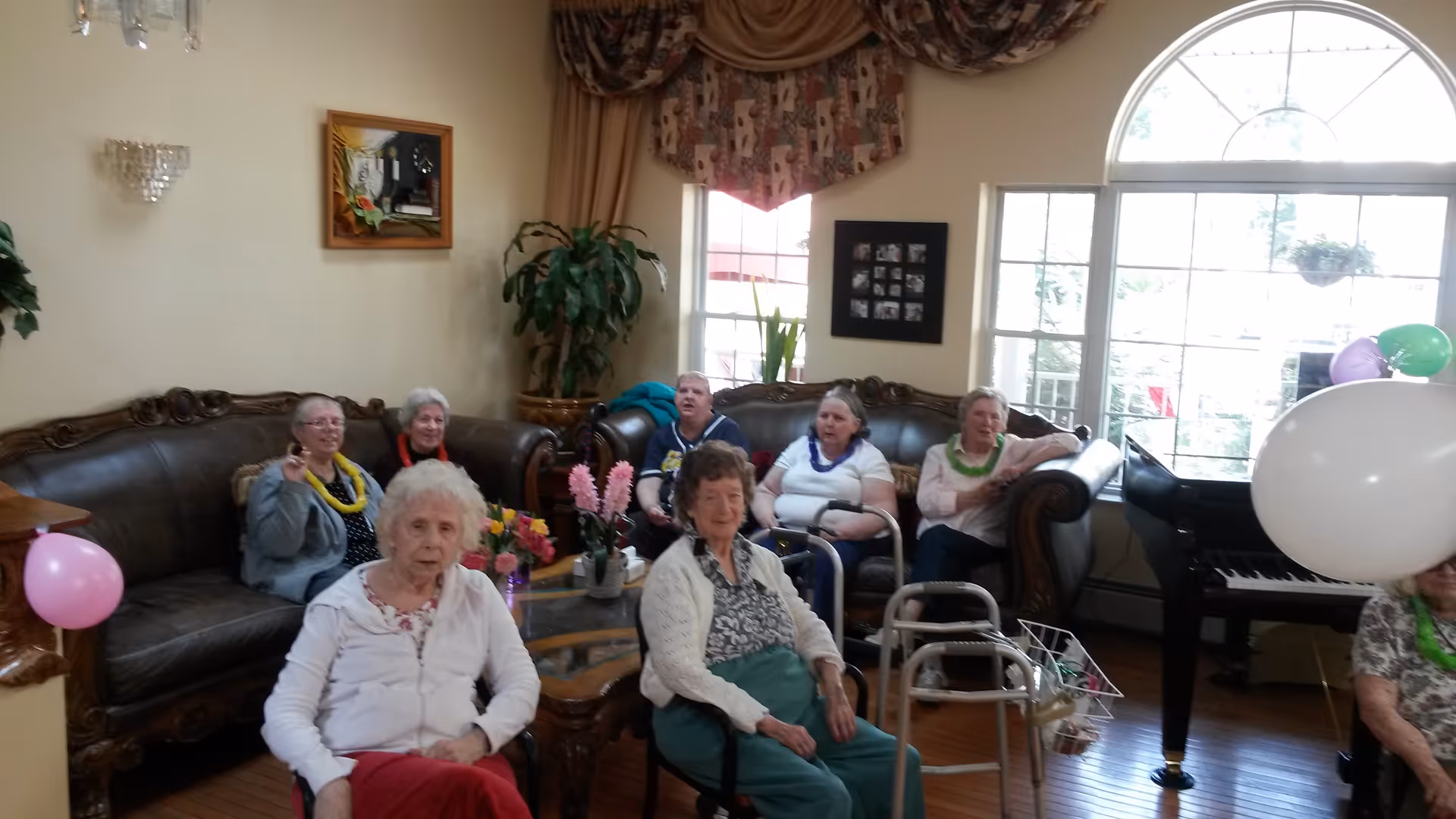 A group of elderly women sitting in a living room with large windows and a grand piano. Some women are seated on leather sofas while others are in chairs. The room is decorated with plants, flowers, balloons, and a framed picture on the wall.