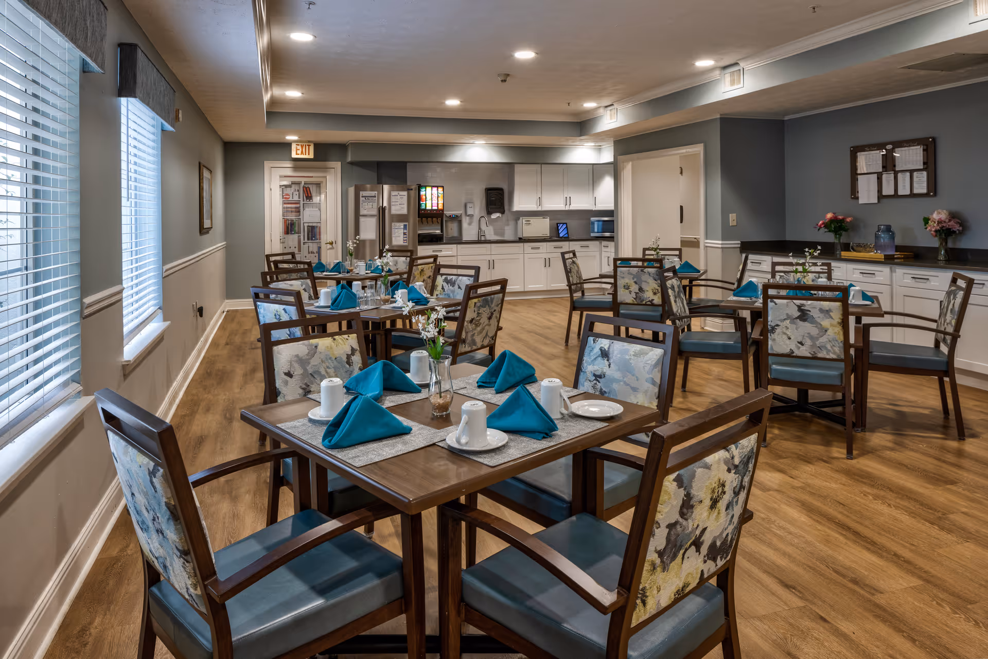A dining room in a senior living facility with several wooden tables and chairs arranged neatly. Each table is set with teal folded napkins, white plates, cups, and small flower vases. The room has wood flooring, light gray walls, and large windows with blinds on the left side. In the background, there is a kitchen area with white cabinets, a soda machine, microwave, and other appliances. The room is well-lit with recessed ceiling lights.