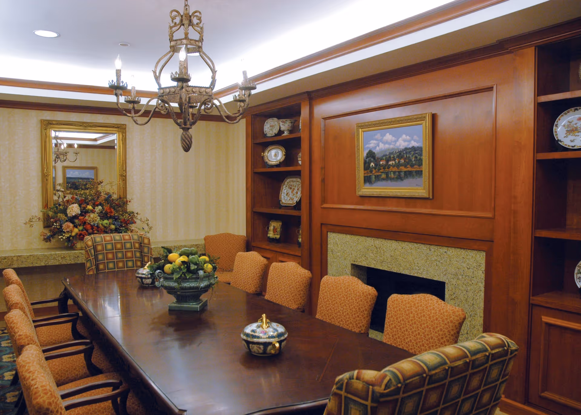 A formal dining room with a long wooden table surrounded by upholstered chairs. The room features a decorative chandelier, built-in wooden shelves with decorative plates, a fireplace with a framed landscape painting above it, and a large mirror reflecting part of the room. There is a floral arrangement on a side table and a fruit centerpiece on the dining table.