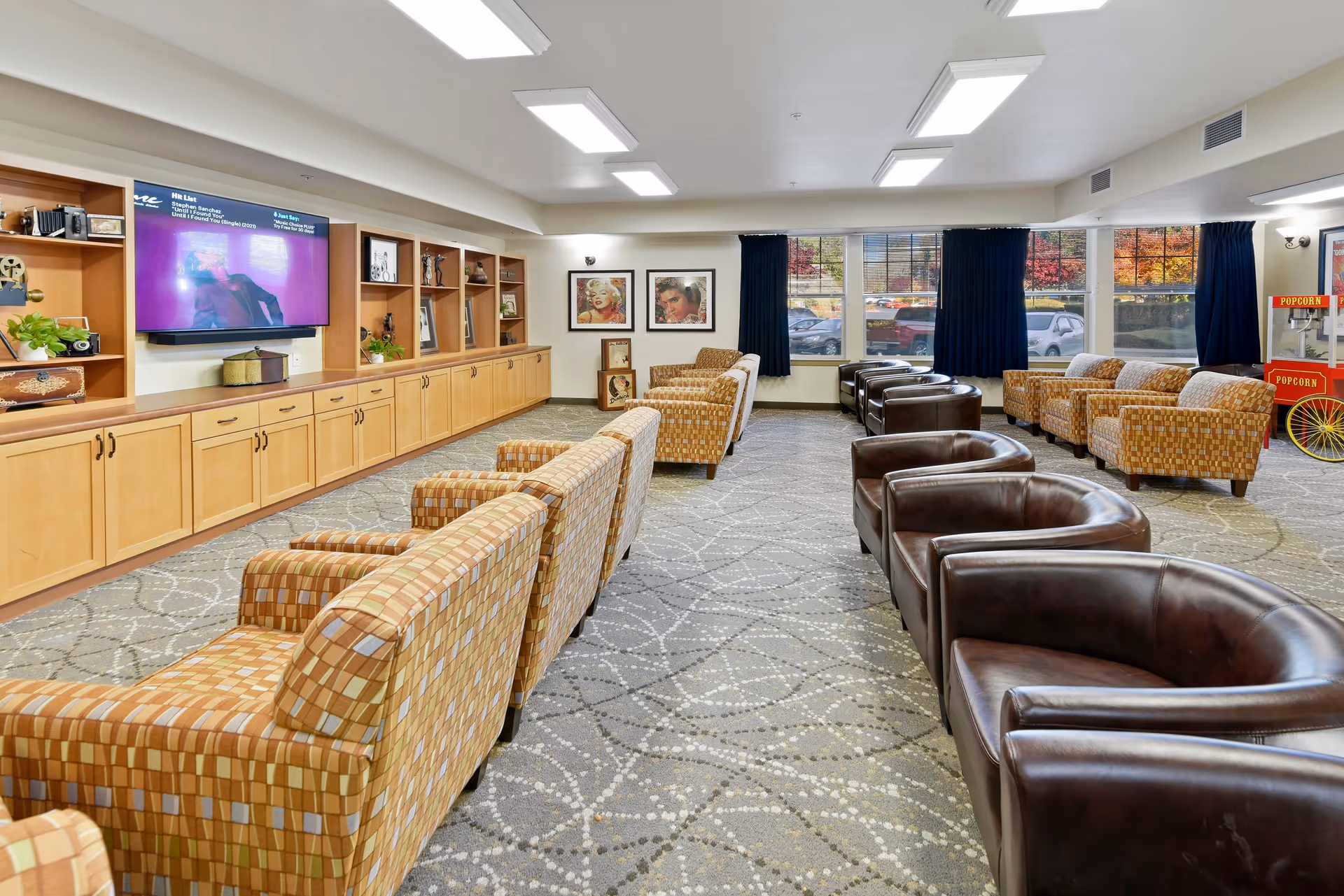 A spacious living room area with rows of patterned armchairs and brown leather chairs facing a large flat-screen TV mounted on a wooden built-in cabinet. The room has large windows with dark curtains, framed artwork on the walls, and a popcorn machine in the corner. The carpet has a geometric pattern and the ceiling has multiple rectangular light fixtures.
