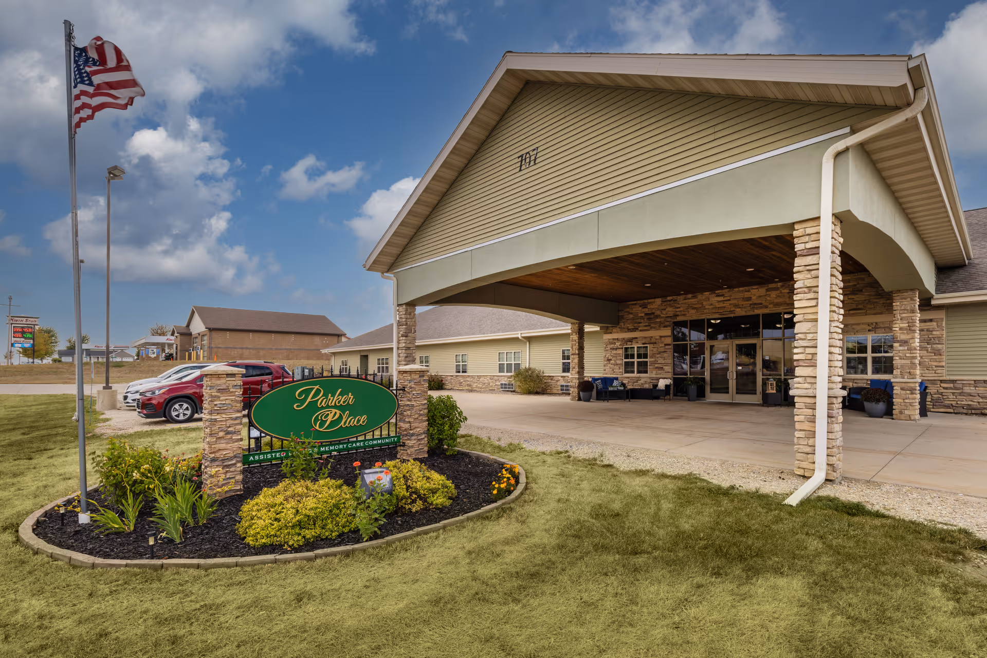 Exterior view of Parker Place Retirement Community building entrance with a covered drop-off area, stone pillars, and a green sign surrounded by landscaping. An American flag is flying on a flagpole to the left, and several parked cars are visible in the background under a partly cloudy sky.
