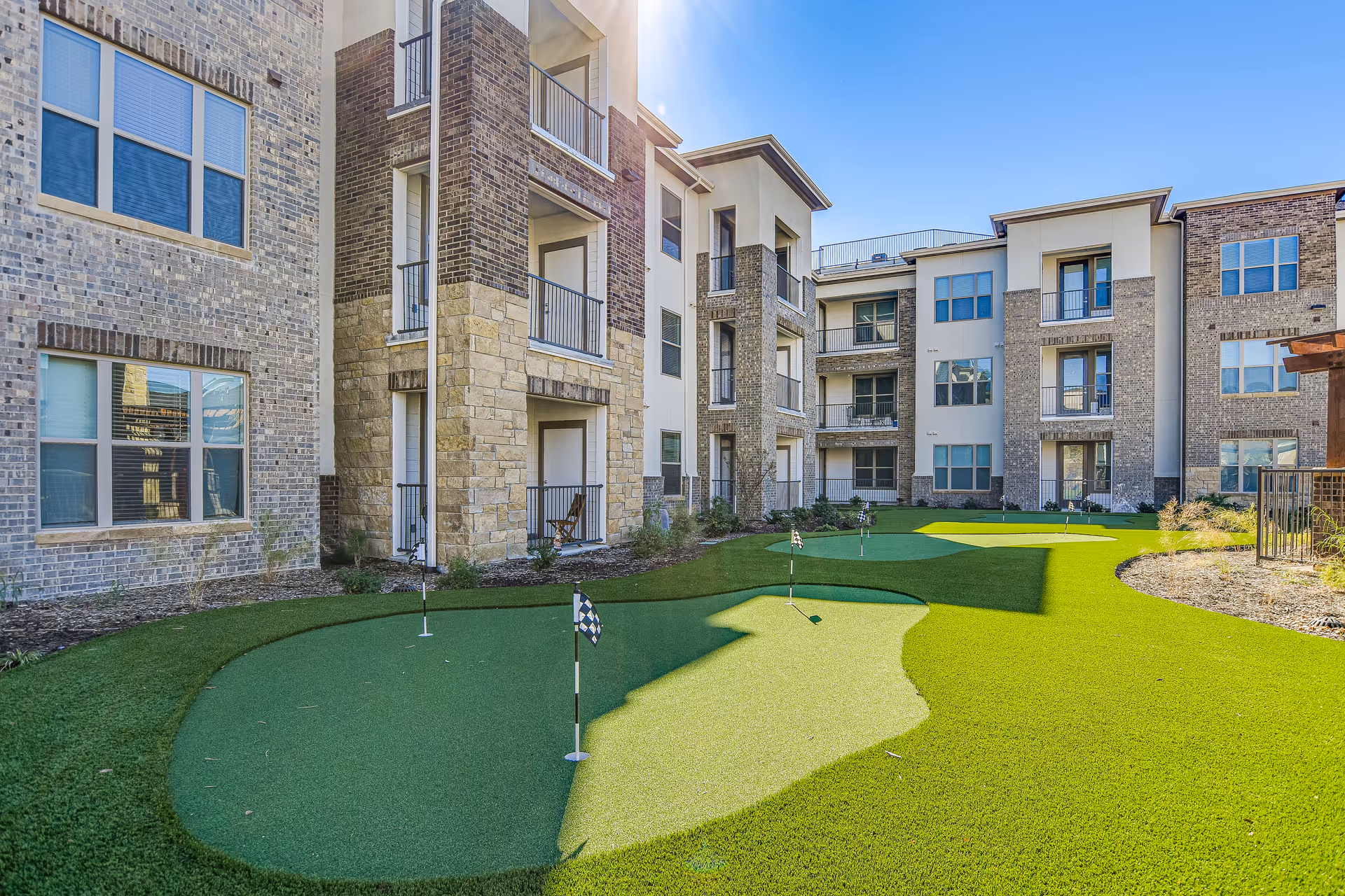 Outdoor courtyard area of a senior living facility featuring a putting green with several golf holes and flags, surrounded by multi-story brick and stone buildings with balconies and windows under a clear blue sky.