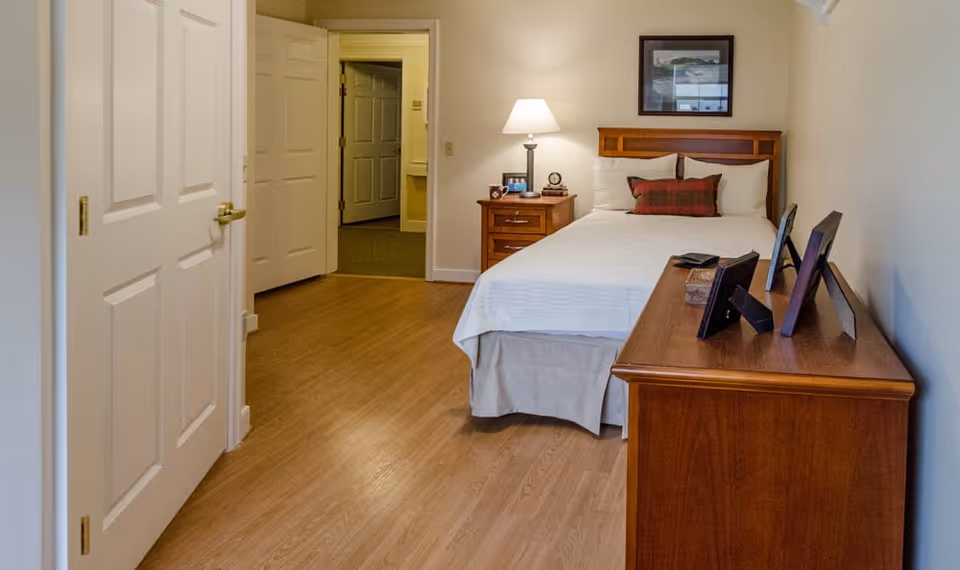 A cozy bedroom in a senior living facility featuring a neatly made bed with white linens and a red plaid pillow, a wooden nightstand with a lamp and framed photos, and a wooden dresser with several framed pictures on top. The room has light-colored walls and wood flooring, with an open door leading to a hallway.
