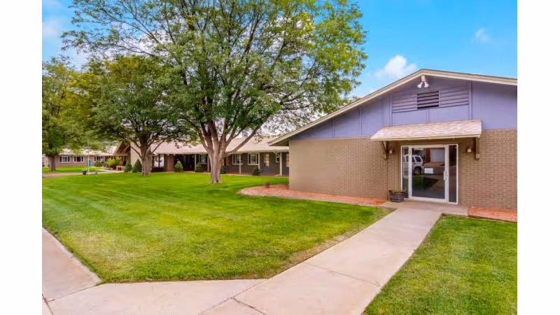 Exterior view of a single-story building with a brick and purple facade, surrounded by a well-maintained green lawn and several large trees under a partly cloudy sky. A concrete walkway leads to a glass door entrance with a small awning above it.