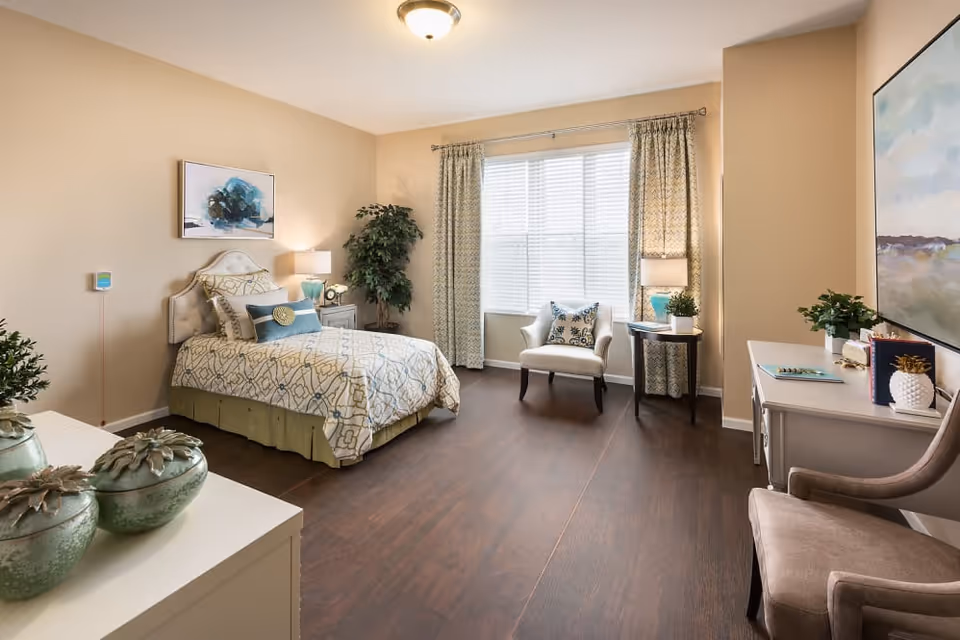 A well-lit bedroom in Cedarbrook of Northville featuring a single bed with patterned bedding, a nightstand with a lamp and clock, a potted plant in the corner, a window with curtains, an armchair with a decorative pillow, a side table with a lamp and plant, a desk with books and decor, and a cushioned chair. The room has wooden flooring and beige walls with artwork hanging.